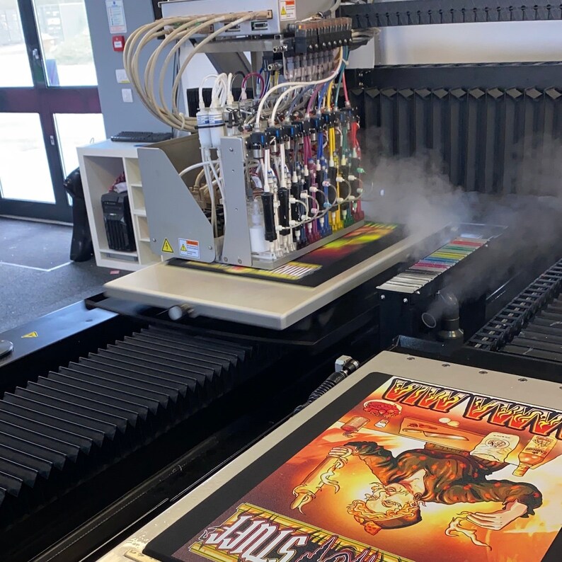 May include: A close-up of a large-format digital printer with a multi-color ink system. The printer is printing a design onto a flat surface. The design features a cartoon character with a skull and crossbones.