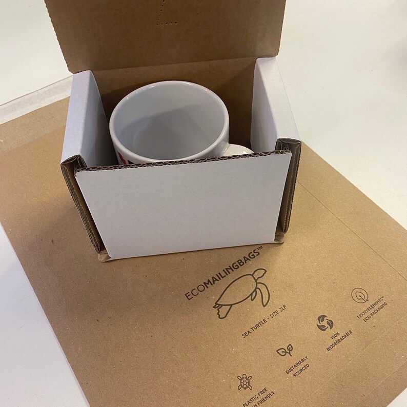 May include: A white ceramic mug with a red and white design inside a brown cardboard box. The box is labeled with the text "ECOMAILINGBAGS" and a sea turtle logo. The box also has a logo with the text "SUSTAINABLY SOURCED" and a recycling symbol.