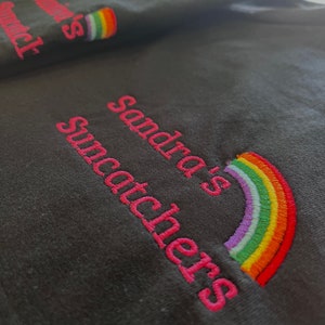 May include: Black t-shirt with pink embroidery that reads "Sandra's Suncatchers" and a rainbow graphic.