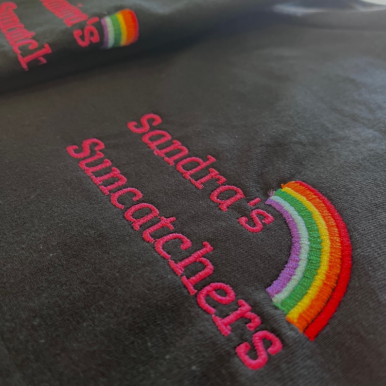 May include: Black t-shirt with pink embroidery that reads "Sandra's Suncatchers" and a rainbow graphic.