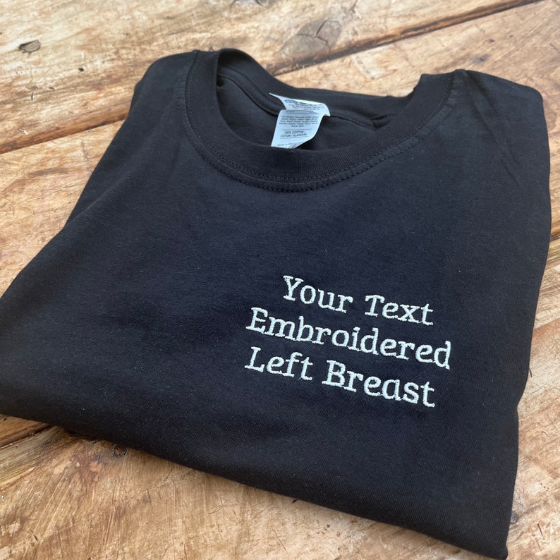 May include: Black t-shirt with white embroidery that says "Your Text Embroidered Left Breast".