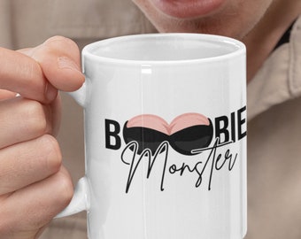 May include: White ceramic coffee mug with a black and pink graphic that says "Boobie Monster".