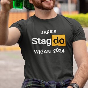 May include: A dark gray t-shirt with white text that reads "JAKE'S Stagdo WIGAN 2024". The word "Stagdo" is in a yellow box.