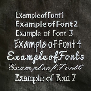 May include: A close-up of a black fabric with seven examples of different fonts in white embroidery. The text reads: "Example of Font 1", "Example of Font 2", "Example of Font 3", "Example of Font 4", "Example of Fonts", "Example of Font 6", and "Example of Font 7".