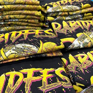 May include: A stack of black t-shirts with a yellow and pink graphic print that says "Rabid" and "Riffes".