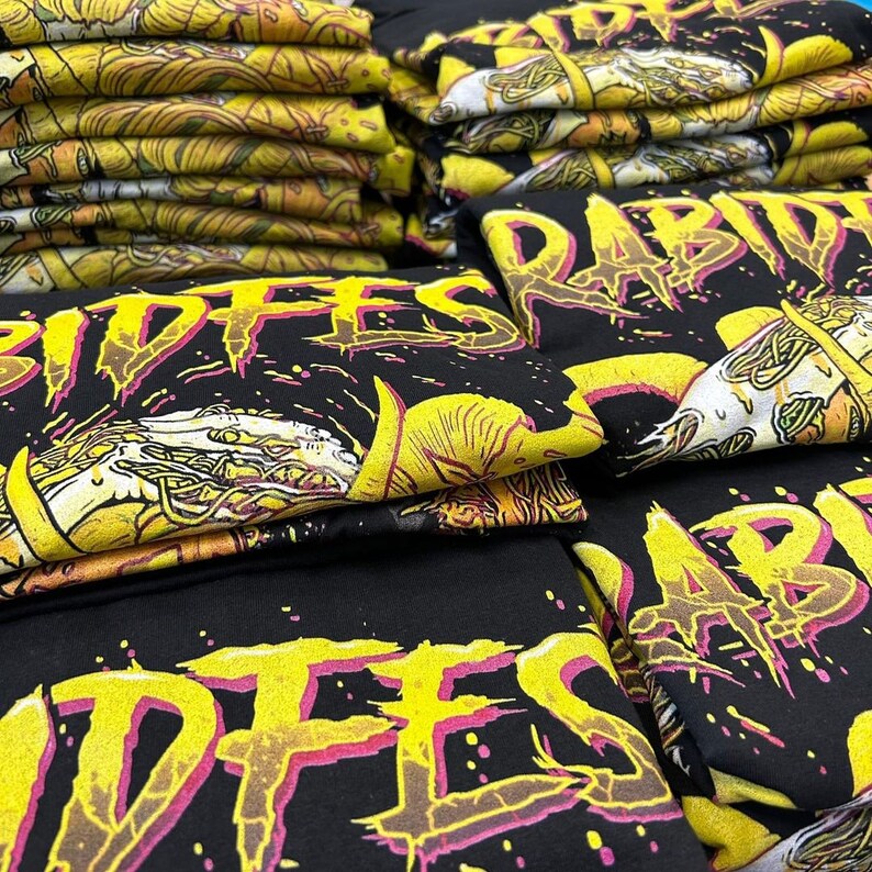 May include: A stack of black t-shirts with a yellow and pink graphic print that says "Rabid" and "Riffes".