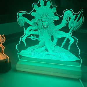 May include: A green and white acrylic lamp with a detailed image of a multi-armed deity with a crown and flowing hair. The deity is sitting on a throne with a floral design.