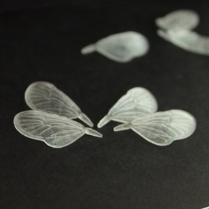 Può includere: Cinque ali di farfalla in plastica trasparente su uno sfondo nero. Le ali sono traslucide e hanno un design delicato e intricato.