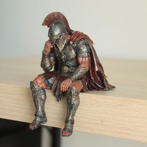 Könnte beinhalten: Eine detaillierte Figur eines sitzenden Kriegers in metallisch silberner Rüstung mit aufwendigen Gravuren, einem dunkelroten Umhang und einem behelmten Kopf. Die Figur ist so positioniert, dass eine Hand das Gesicht berührt, mit realistischen Muskeln und verwitterten Texturen.