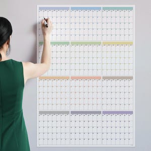 May include: A large white wall calendar with a grid layout, divided into sections with pastel-colored headers. A person is seen writing on the calendar with a black marker. The calendar is mounted on a light gray wall.