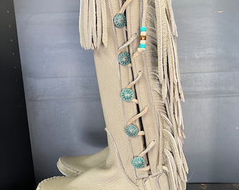 May include: Beige leather knee-high boots with fringe detailing and turquoise button accents. The boots have a moccasin-style toe and are laced up the side. The boots are hanging from a black hanger.