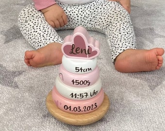 以下が含まれることがあります： 「Leni」と刻まれたピンクと白の積み木のおもちゃ。リングには赤ちゃんの出生情報も記載されています。51cm、4500g、11:57、01.03.2023。小さな女の子がおもちゃの前に座っています。