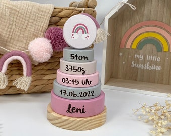 May include: A wooden stacking toy with a rainbow design on the top piece. The other pieces are pink and gray and have the following information written on them: 51cm, 3750g, 03:45 Uhr, 17.06.2022, Leni.