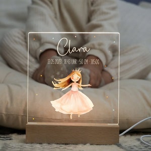 Könnte beinhalten: Ein klares Acryl-Nachtlicht mit einem Holzsockel. Das Licht zeigt eine Prinzessinnen-Illustration mit einer Krone, einem rosa Kleid und blonden Haaren. Der Text "Clara" und "13.05.2023 - 18:43 UHR - 50 CM - 3850G" ist auf das Licht gedruckt.