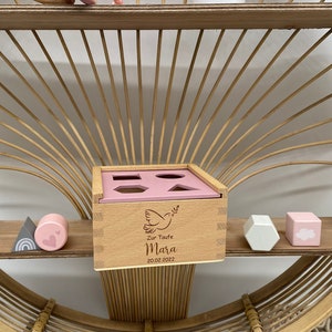 May include: Wooden shape sorter toy with a pink lid and various geometric cutouts. The wooden box is engraved with a dove, the name "Mara", and the date "20.02.2022". Additional wooden shapes are also visible.