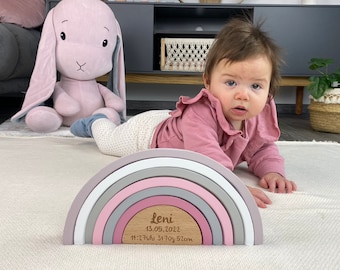 May include: A wooden rainbow stacking toy with a personalized plaque that reads "Leni 13.05.2022 14:27 Uhr 3470g 52cm". The rainbow is made of five different colored arches, with the top arch being white, followed by pink, gray, pink, and gray. A baby is playing with the toy.