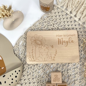 May include: A wooden engraved board with the text "Hier kr&uuml;melt Mayla" and a small bunny illustration. The board is surrounded by a beige macrame and a few wooden blocks with a heart, a cloud, and the letters A, B, and C.