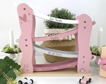 May include: Pink wooden toy race track with three lanes and a white base. The track has a white and gray ramp with the text "Zur Taufe" and "Malia" and the date "15.10.2022". There are four small toy cars, two pink and two white, on a white surface.