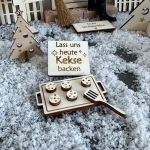 Könnte beinhalten: Holz Schild mit dem Text "Lass uns heute Kekse backen". Das Schild liegt auf einer weißen Oberfläche mit einer Holz Backform mit fünf Keksen und einem Spatel.
