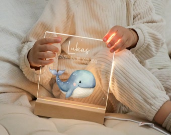 May include: A personalised night light with a blue whale design and the name "Lukas" in script. The light is made of clear acrylic and sits on a wooden base. The light is plugged in with a white cord.