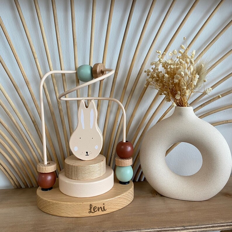 May include: Wooden toy with a bunny face and colorful beads on a track. The toy is on a wooden base with the name "Leni" engraved on it.
