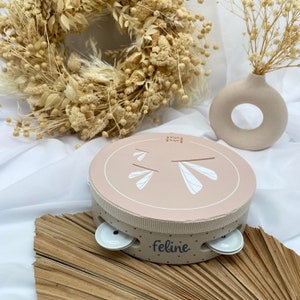 Può includere: Un tamburello rotondo rosa chiaro con un disegno di libellula e la scritta "Feline" stampata sul lato. Il tamburello ha piccoli piatti bianchi e una fascia in tessuto fantasia. La parte superiore presenta un disegno di libellula.