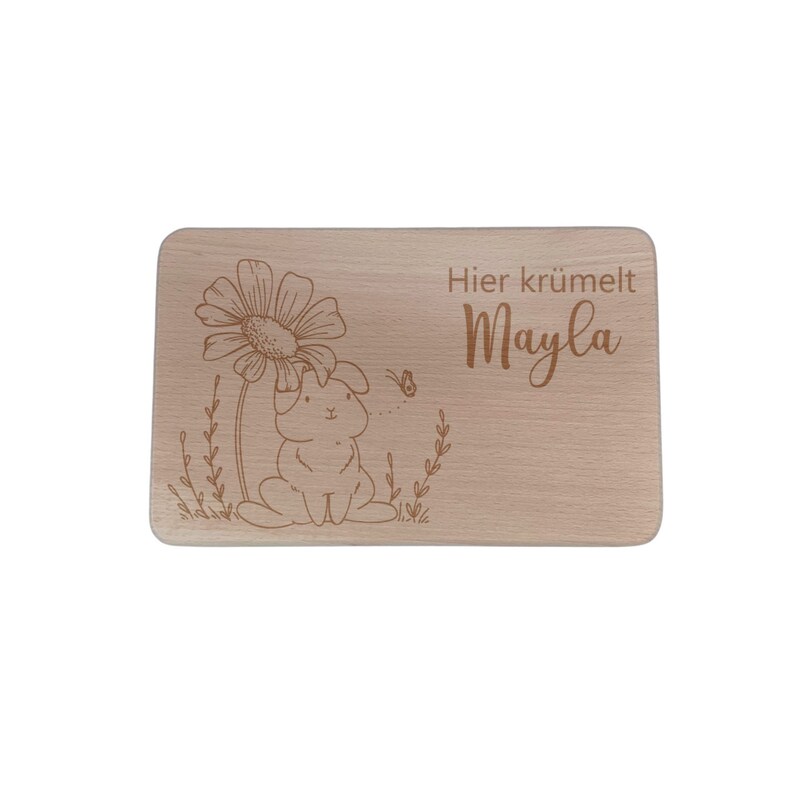 May include: A wooden cutting board with a laser-engraved design of a bunny rabbit sitting under a flower. The text "Hier kr&uuml;melt Mayla" is engraved on the board.