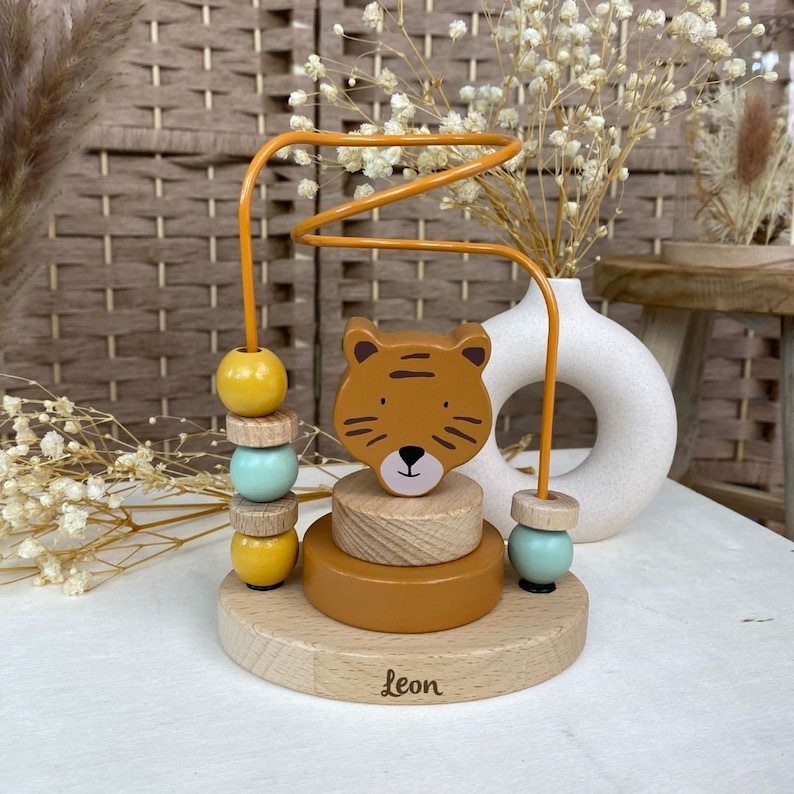 May include: A wooden toy with a tiger face and a winding wire for beads. The toy has a base with the name "Leon" written on it. The beads are yellow and blue.