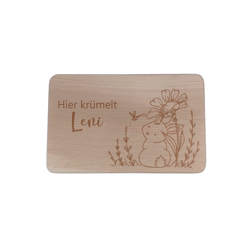 May include: A wooden cutting board with a laser-engraved design of a bunny rabbit sitting in a field of flowers. The text "Hier kr&uuml;melt Leni" is also engraved on the board.
