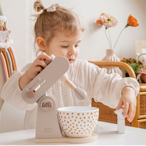 May include: A child plays with a wooden toy stand mixer. The mixer is light gray with a brown base and a white bowl with brown polka dots. The mixer has a white handle and a white whisk attachment. The mixer has a label that says "Lilli" on it.