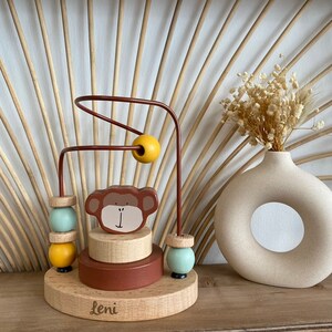 May include: Wooden toy with a monkey face, colorful beads, and a wire maze. The base of the toy is brown and has the name "Leni" written on it.