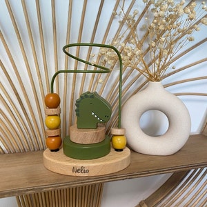 May include: A wooden toy with a green dinosaur head and a colorful bead maze. The toy is on a wooden base with the name "Nelio" engraved on it.