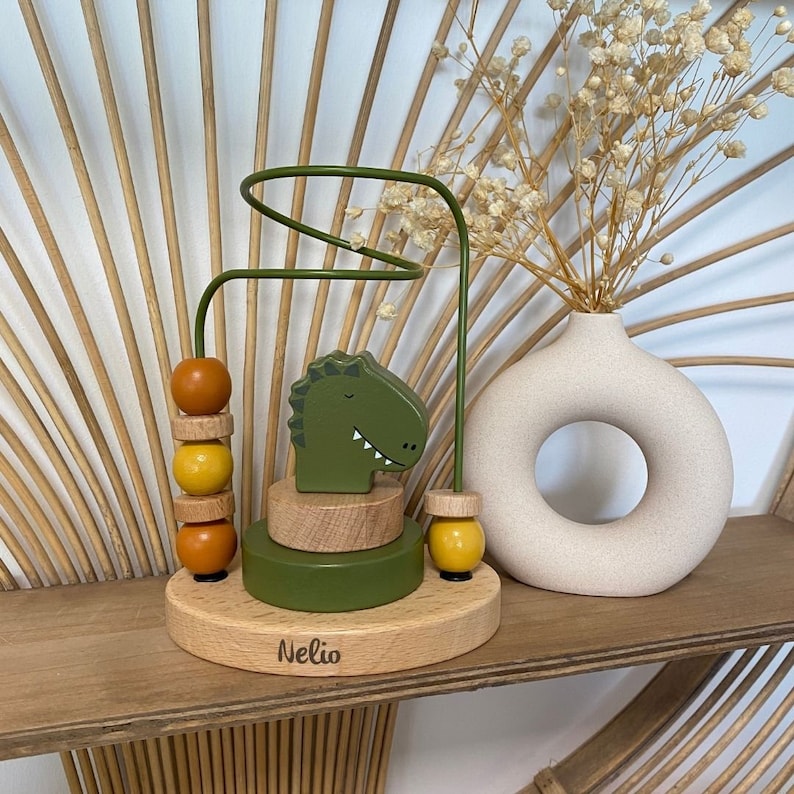 May include: A wooden toy with a green dinosaur head and a colorful bead maze. The toy is on a wooden base with the name "Nelio" engraved on it.