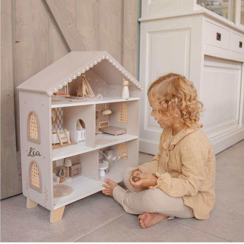 Puede incluir: Una casa de mu&ntilde;ecas de madera de tres pisos en tonos neutros con techo a dos aguas. La casa de mu&ntilde;ecas presenta cortes decorativos, un nombre en el lateral y muebles en miniatura. La casa de mu&ntilde;ecas est&aacute; sobre patas de madera.