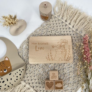 May include: A wooden cutting board with the text "Hier kr&uuml;melt Leni" engraved on it. The board is surrounded by a beige crocheted blanket, a beige silicone bib with a leopard print, and wooden blocks with letters and a heart and cloud design.