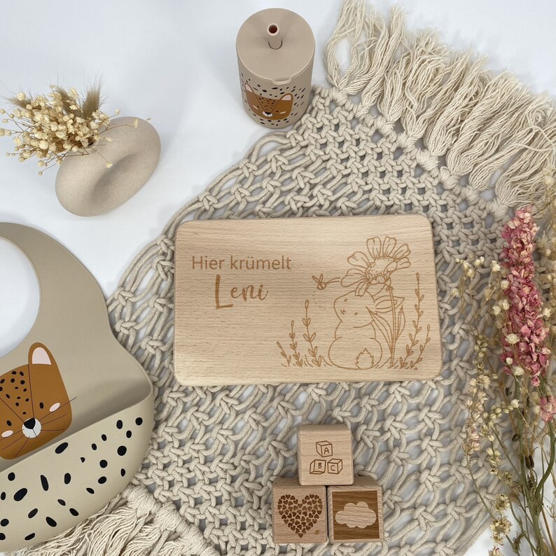 May include: A wooden cutting board with the text "Hier kr&uuml;melt Leni" engraved on it. The board is surrounded by a beige crocheted blanket, a beige silicone bib with a leopard print, and wooden blocks with letters and a heart and cloud design.