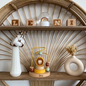 May include: A wooden toy with a lion face and colorful beads on a yellow track. The toy is on a wooden shelf with a snow globe and wooden blocks that spell "LENI".