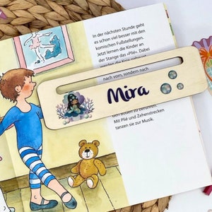 Puede incluir: Un marcapáginas de madera con una ilustración de sirena y el nombre "Mira" impreso en morado. El marcapáginas tiene una ranura para sujetar una página y presenta un acabado en madera clara.