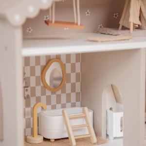 Puede incluir: Una casa de mu&ntilde;ecas de madera de varios niveles con una paleta de colores neutros. Cuenta con un columpio, un tipi en miniatura, un ba&ntilde;o con ba&ntilde;era, espejo y escalera, y una peque&ntilde;a cuna. Las paredes est&aacute;n decoradas con un patr&oacute;n de cuadros y detalles florales.