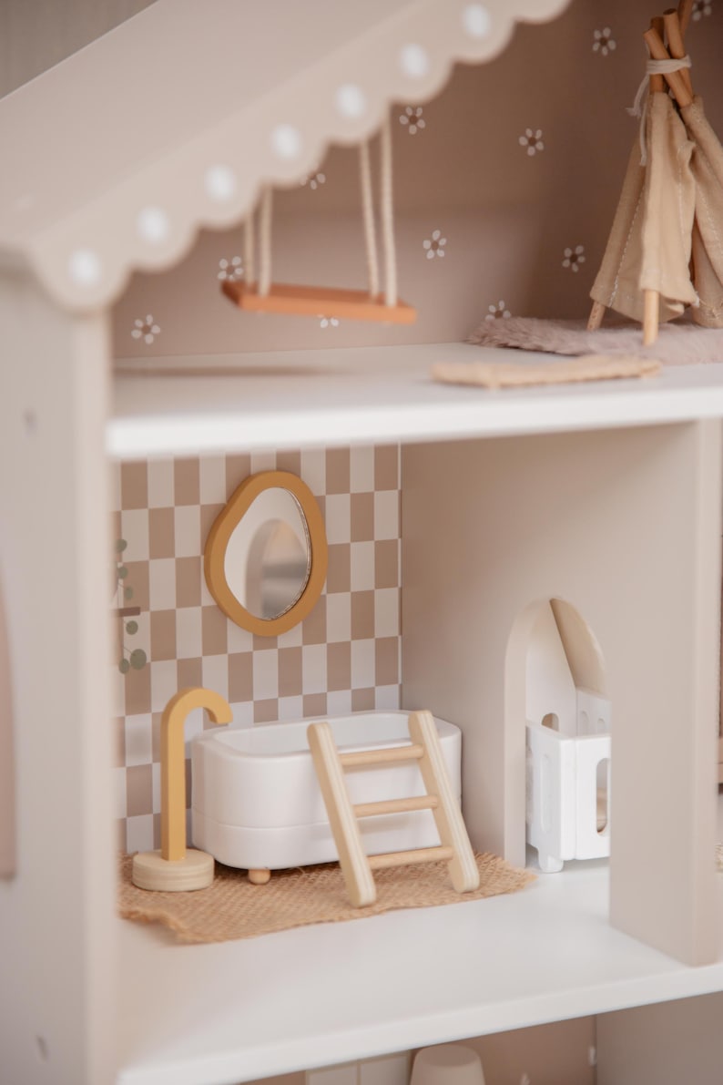 Puede incluir: Una casa de mu&ntilde;ecas de madera de varios niveles con una paleta de colores neutros. Cuenta con un columpio, un tipi en miniatura, un ba&ntilde;o con ba&ntilde;era, espejo y escalera, y una peque&ntilde;a cuna. Las paredes est&aacute;n decoradas con un patr&oacute;n de cuadros y detalles florales.
