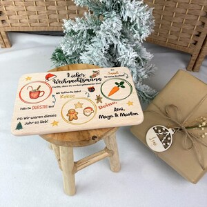 May include: A wooden Christmas sign with a festive design. The sign says "Lieber Weihnachtsmann" and includes a mug of hot chocolate, gingerbread cookies, a carrot, and a message for Santa. The sign is decorated with stars and a red Santa hat.