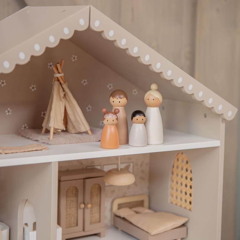 Puede incluir: Una casa de mu&ntilde;ecas de madera beige con un techo festoneado y detalles blancos. En el interior, hay un tipi en miniatura, una familia de mu&ntilde;ecos de madera y habitaciones amuebladas. La casa de mu&ntilde;ecas presenta un dormitorio, un armario y una ventana decorativa.
