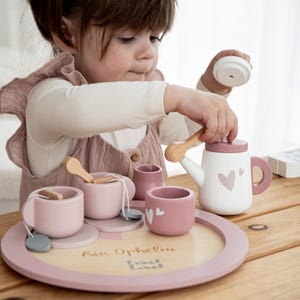 Può includere: Un set da tè in legno rosa e bianco, composto da una teiera con dettagli a cuore, tazze, piattini e cucchiaini. Il set è presentato su un vassoio rotondo con la scritta "Ria Ophelia".