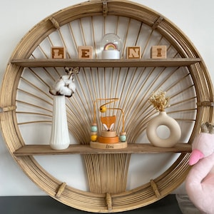 May include: A round, woven shelf with two shelves. The top shelf has a snow globe, three wooden blocks spelling "LENI", and a wooden fox toy. The bottom shelf has a white vase with cotton flowers, a wooden fox toy, and a white vase with dried flowers.