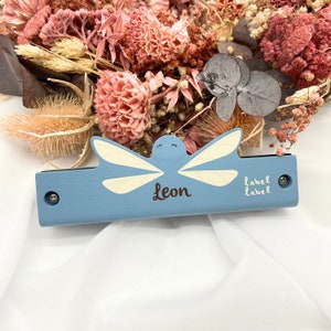May include: A light blue wooden nameplate with a dragonfly design and the name "Leon" in brown script. The dragonfly has cream-colored wings. The nameplate is decorated with two screws and the words "label label".