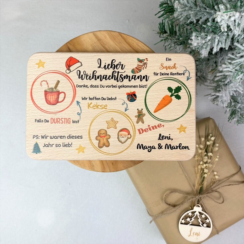 May include: A wooden sign with a Christmas theme. It says "Lieber Weihnachtsmann" and has illustrations of a mug of hot chocolate, cookies, a carrot, and Santa Claus. The sign also says "Danke, dass Du vorbei gekommen bist!" and "Wir hoffen Du liebst Kekse". It is signed "Deine, Leni, Maya & Marlon".