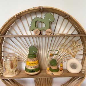 May include: Three wooden dinosaur toys for a baby. The first toy is a pull toy with a green dinosaur and the text "Born 07.06.2023 03:45Uhr 3750g Nelio". The second toy is a stacking toy with green and yellow rings and the text "5cm 3750g 03:45Uhr 17.06.2023 Nelio". The third toy is a green dinosaur with a yellow bead and the text "Nelio".