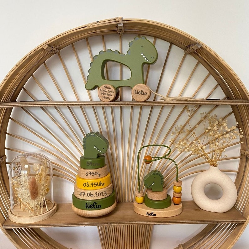 May include: Three wooden dinosaur toys for a baby. The first toy is a pull toy with a green dinosaur and the text "Born 07.06.2023 03:45Uhr 3750g Nelio". The second toy is a stacking toy with green and yellow rings and the text "5cm 3750g 03:45Uhr 17.06.2023 Nelio". The third toy is a green dinosaur with a yellow bead and the text "Nelio".