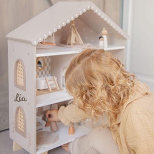 Puede incluir: Una casa de mu&ntilde;ecas de madera beige claro con un techo festoneado y recortes decorativos. La casa de mu&ntilde;ecas tiene varios estantes con muebles y accesorios en miniatura. El nombre "Lia" est&aacute; impreso en el lateral. Un ni&ntilde;o est&aacute; jugando con la casa de mu&ntilde;ecas.
