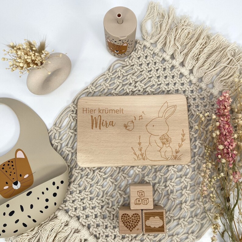 May include: A wooden cutting board with a bunny rabbit illustration and the text "Hier kr&uuml;melt Mira". The board is surrounded by a beige crocheted blanket, a beige silicone bib with a leopard print, and a beige cup with a leopard print. There are also three wooden blocks with a heart, a cloud, and letters.